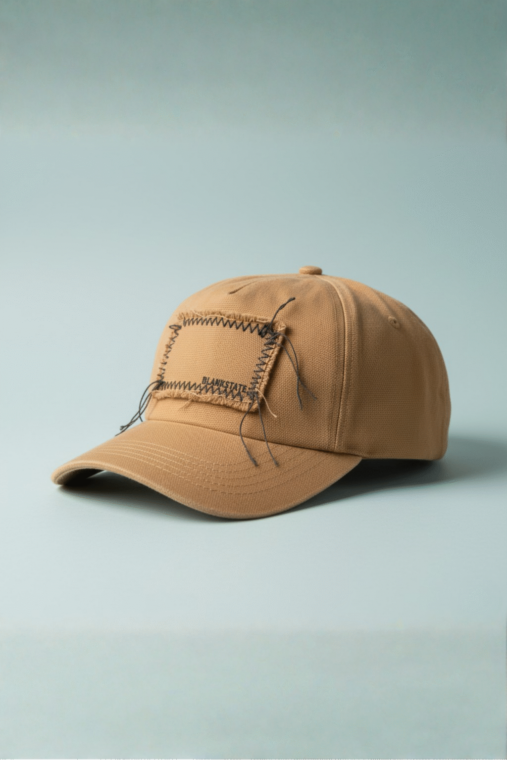 Camel raw-edge canvas cap with stitched rectangular patch on pale bluish background